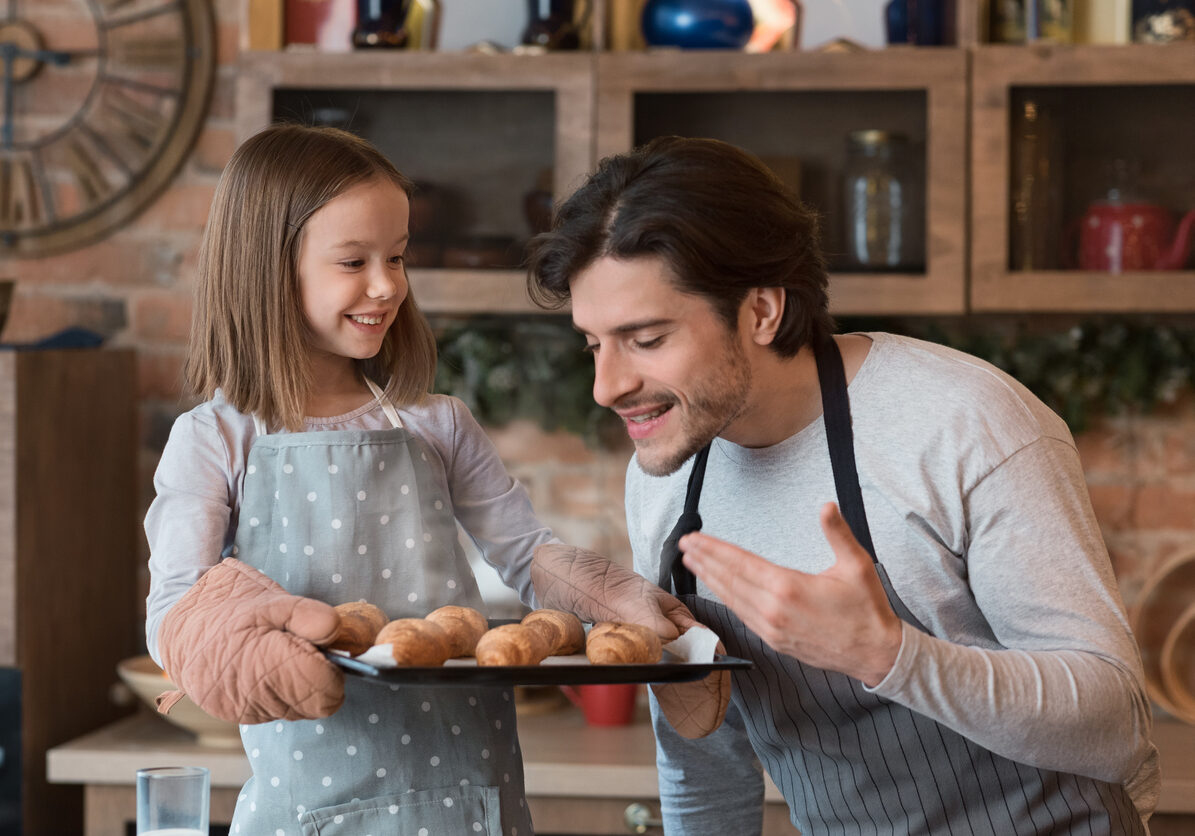 Cute little girl holding tray with fresh baked croissants, happy dad enjoying smell of homemade pastry in kitchen, closeup